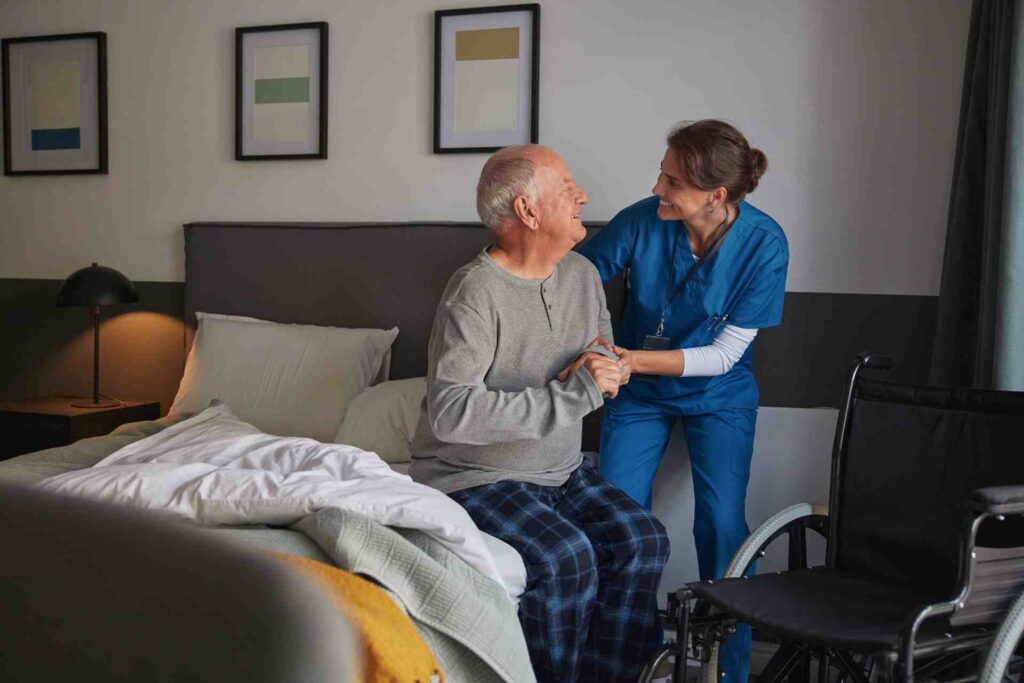 Home health aide helping an older man beside his bed at home